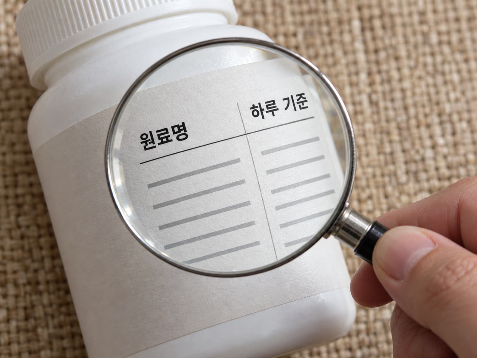 보충제 포장 뒷면의 표시를 돋보기로 확인하는 손.